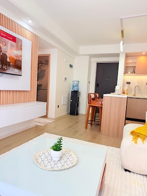 A modern living room with a flat-screen TV, a coffee table with a plant, a sofa with a yellow throw, and a kitchen area with a water dispenser. - 2-Bedroom Westlands Two Balconies Next To Foodplus And Artcaffe Rhapta Road (Nairobi)