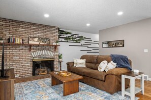 A living room with a brick fireplace, a brown leather sofa, a wooden coffee table, and a patterned rug. - 4 min to Hospitals • Modern 2 BR Stay (Billings)