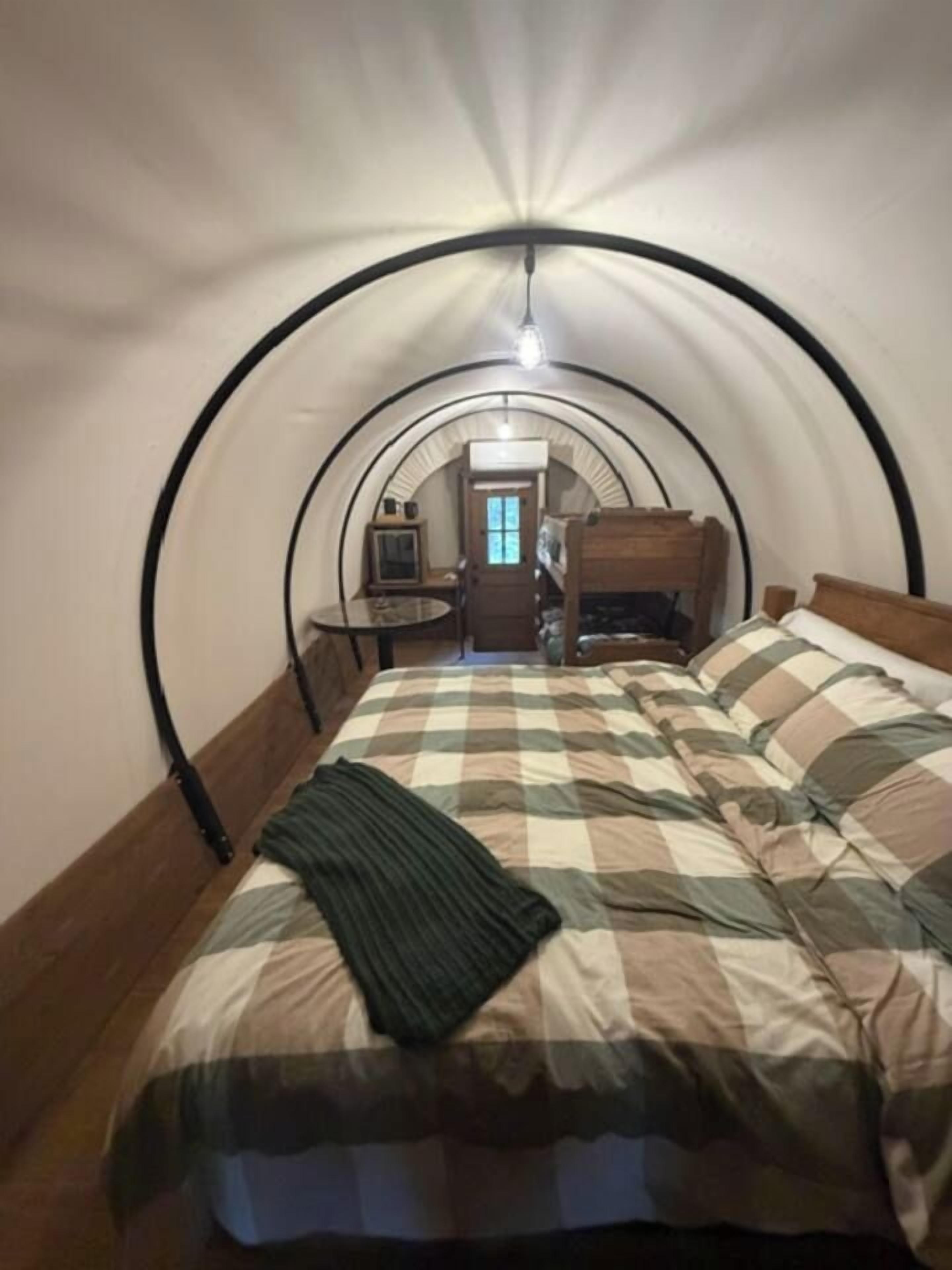 A room with a curved ceiling, a bed with a checkered bedspread, a wooden headboard, and a small table with a chair.
