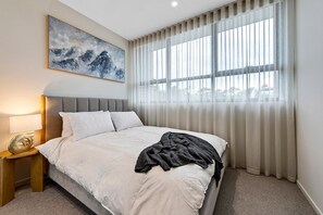 A bedroom with a bed, a nightstand, a lamp, a painting, and a window with curtains. - Spacious 3-bed Townhouse In Mile End (Adelaide)
