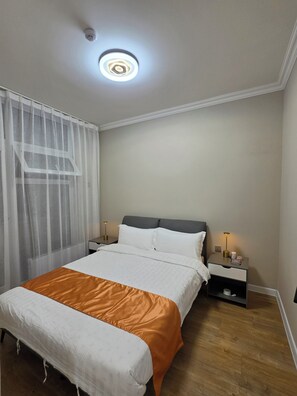 Ett hotellrum med en säng, sängbord, en taklampa och ett fönster med tunna, genomskinliga gardiner.