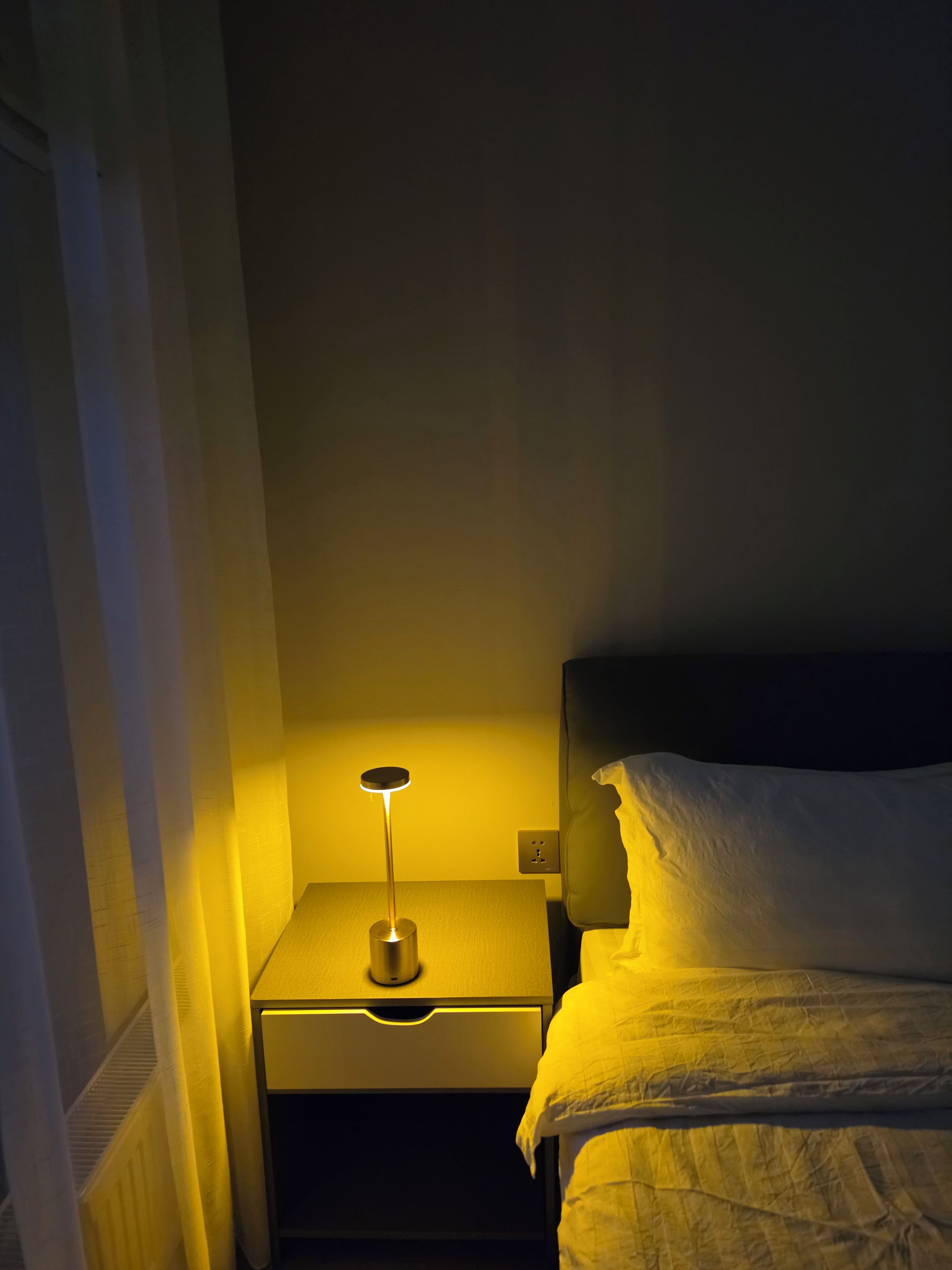 Una mesita de noche pequeña con una lámpara, una cama con ropa blanca y un enchufe de pared.