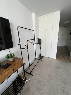 A room with a TV mounted on the wall, a wooden desk with a plant and a power strip, and a black metal rack with a vacuum cleaner. - Ilikai Marina condo for rent with free parking (Honolulu)