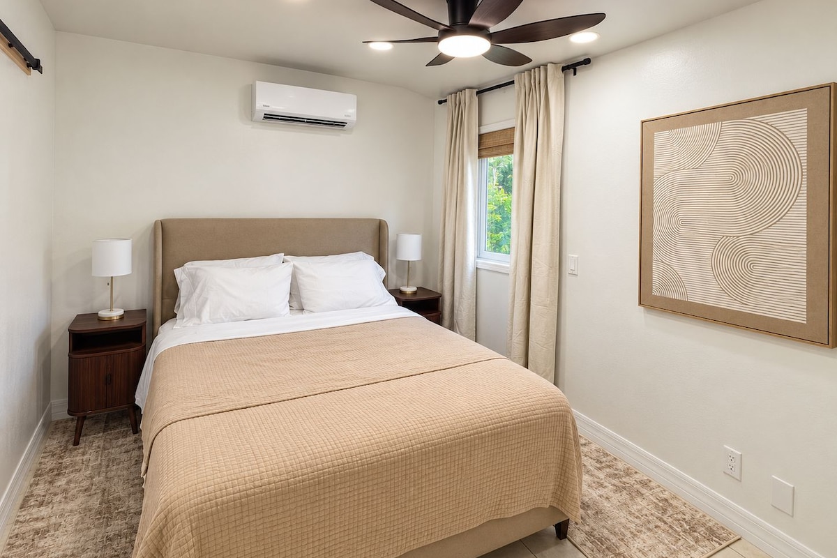 A bedroom with a bed, two bedside tables, a ceiling fan, an air conditioning unit, a window with curtains, and a wall-mounted artwork.