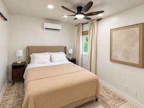 A bedroom with a bed, two bedside tables, a ceiling fan, an air conditioning unit, a window with curtains, and a wall-mounted artwork. - Aqua Suites (Nassau)
