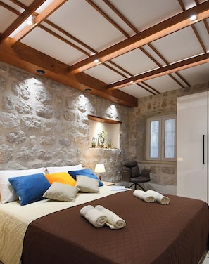 A bedroom with a stone wall, a bed with a brown bedspread, a chair, and a small table with a lamp. - Perfect 1-bedroom apartment with ACin Dubrovnik,quiet Street, steps from Stradun (Dubrovnik)