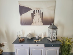 A wall-mounted painting of a pier extending into a calm body of water, a wooden cabinet with drawers, and a decorative lantern. - Cozy Vacation Rental - Porte de Chambord - Garden - Pets Allowed (Huisseau-sur-Cosson)