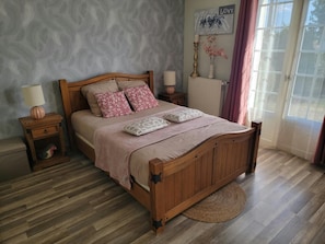 A bedroom with a wooden bed, two bedside tables, a radiator, a wall-mounted artwork, and a window with curtains. - Cozy Vacation Rental - Porte de Chambord - Garden - Pets Allowed (Huisseau-sur-Cosson)