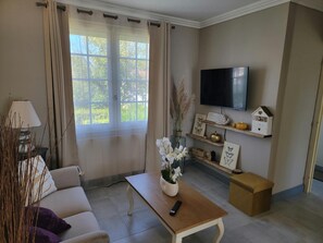 A living room with a sofa, a coffee table with a vase of flowers, a television mounted on the wall, and a window with curtains. - Cozy Vacation Rental - Porte de Chambord - Garden - Pets Allowed (Huisseau-sur-Cosson)