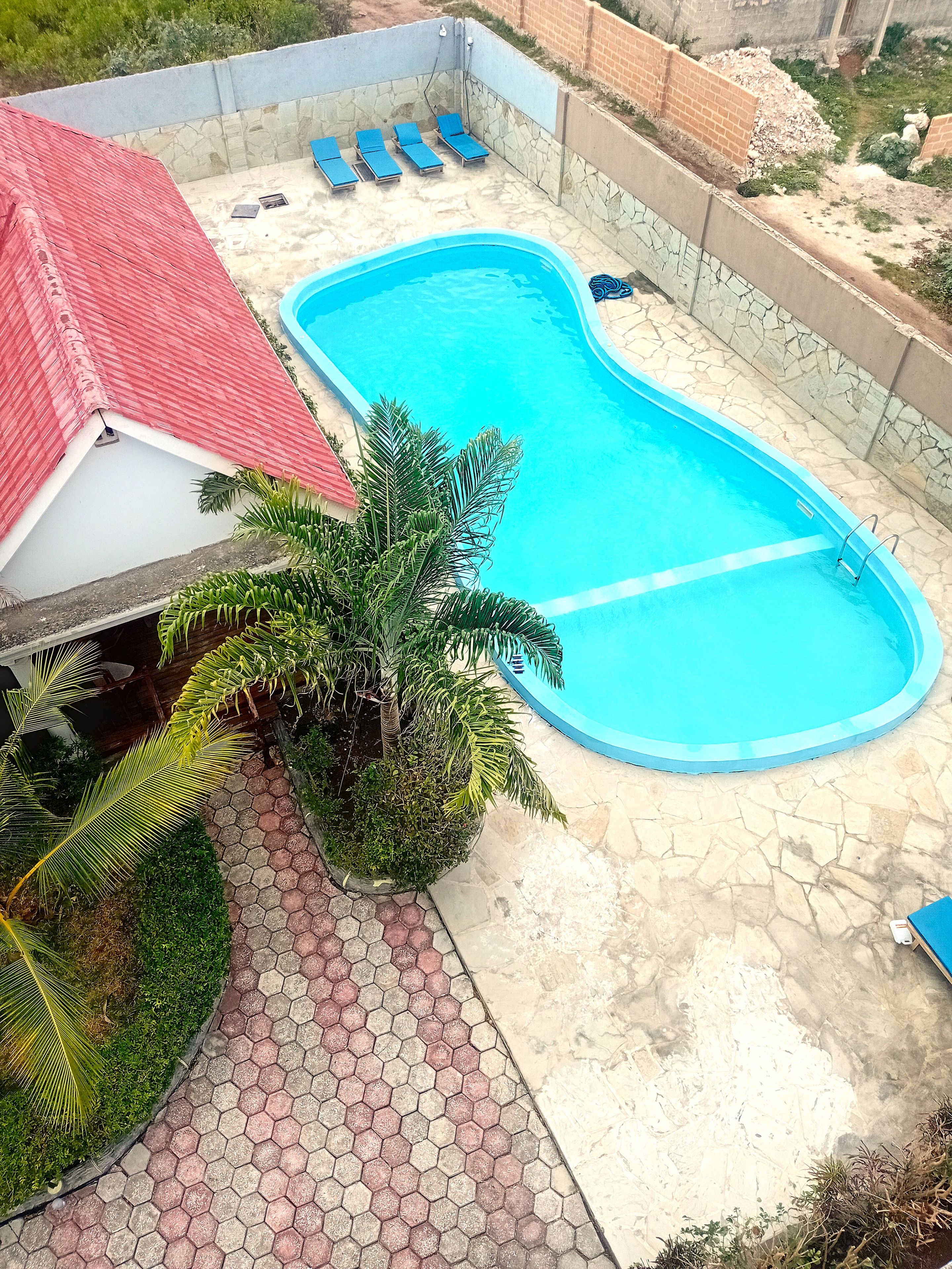 Una piscina con un edificio de techo rojo, un paseo con baldosas hexagonales y una palmera.