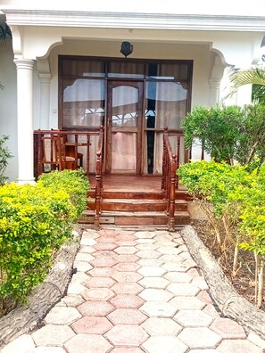 Entrada de un edificio con una escalera de madera que conduce a una puerta, flanqueada por follaje y un camino empedrado.