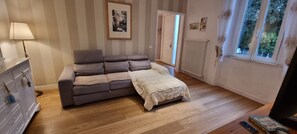 A room with a grey sofa, a white bedside table, a lamp, a framed picture on the wall, a radiator, and a window with a view of trees. - Apartment near the beach (Marina di Pietrasanta)