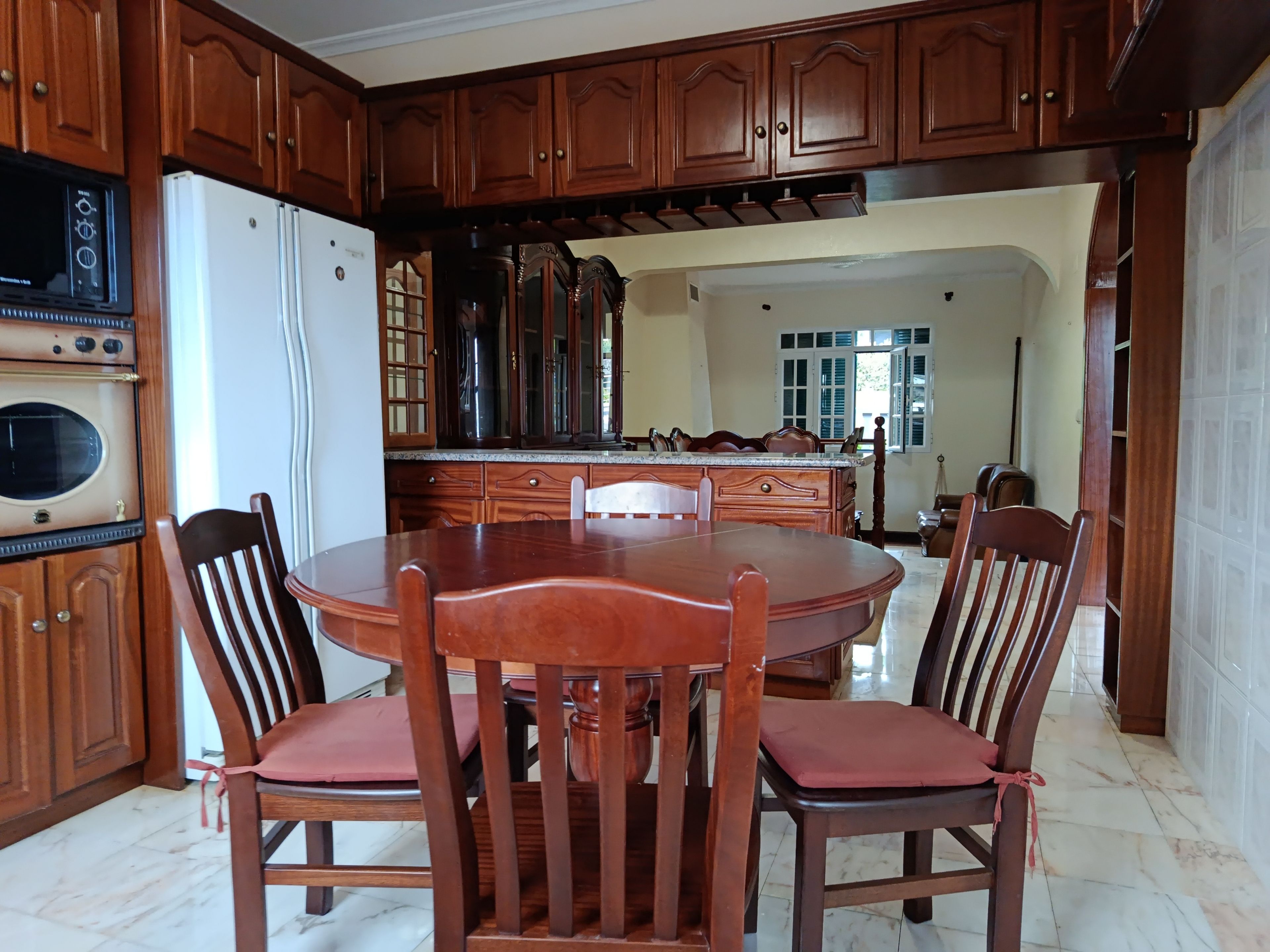 A dining area with a wooden table and chairs, a built-in oven, and a kitchen with wooden cabinets and a refrigerator.
