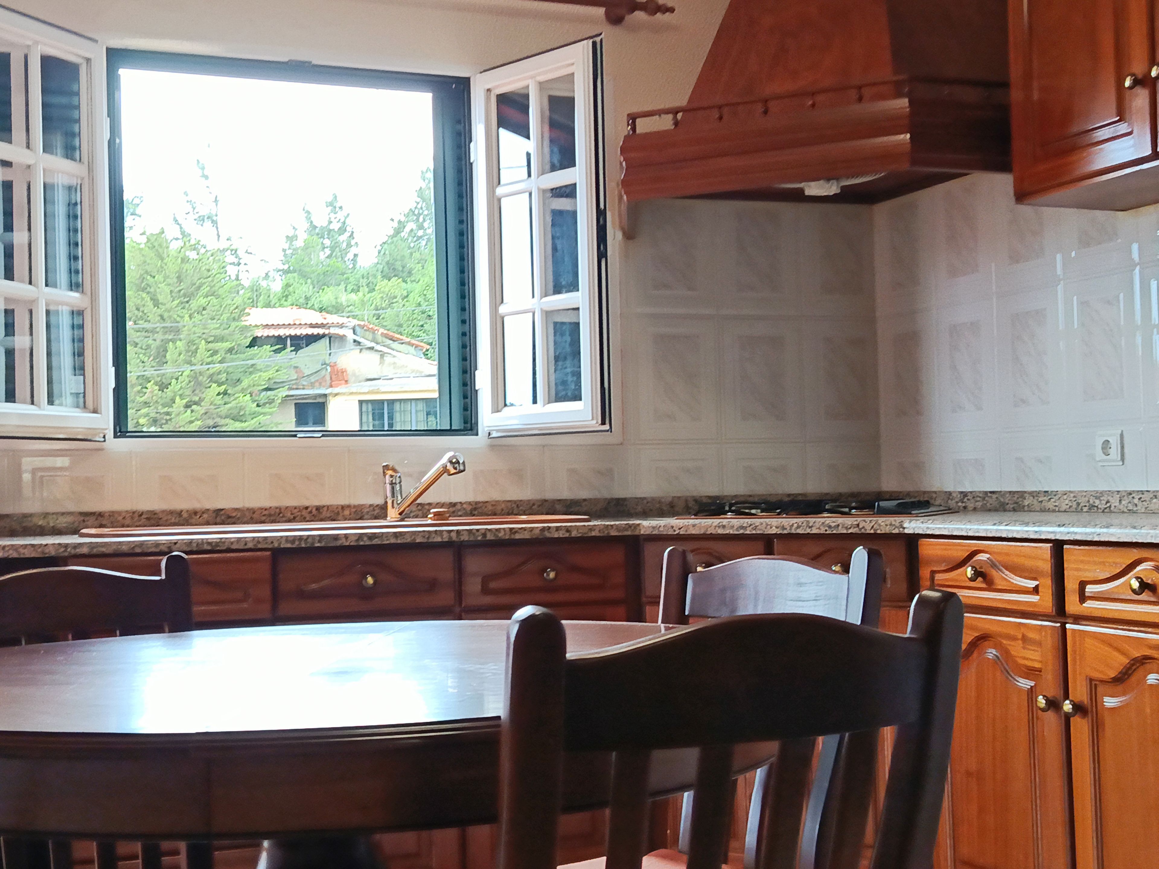 A kitchen with a dining table, chairs, wooden cabinets, a window with open shutters, and a view of greenery outside.