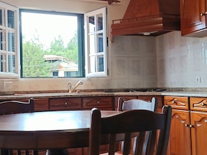 A kitchen with a dining table, chairs, wooden cabinets, a window with open shutters, and a view of greenery outside. - Mist Haven (Santo António da Serra)