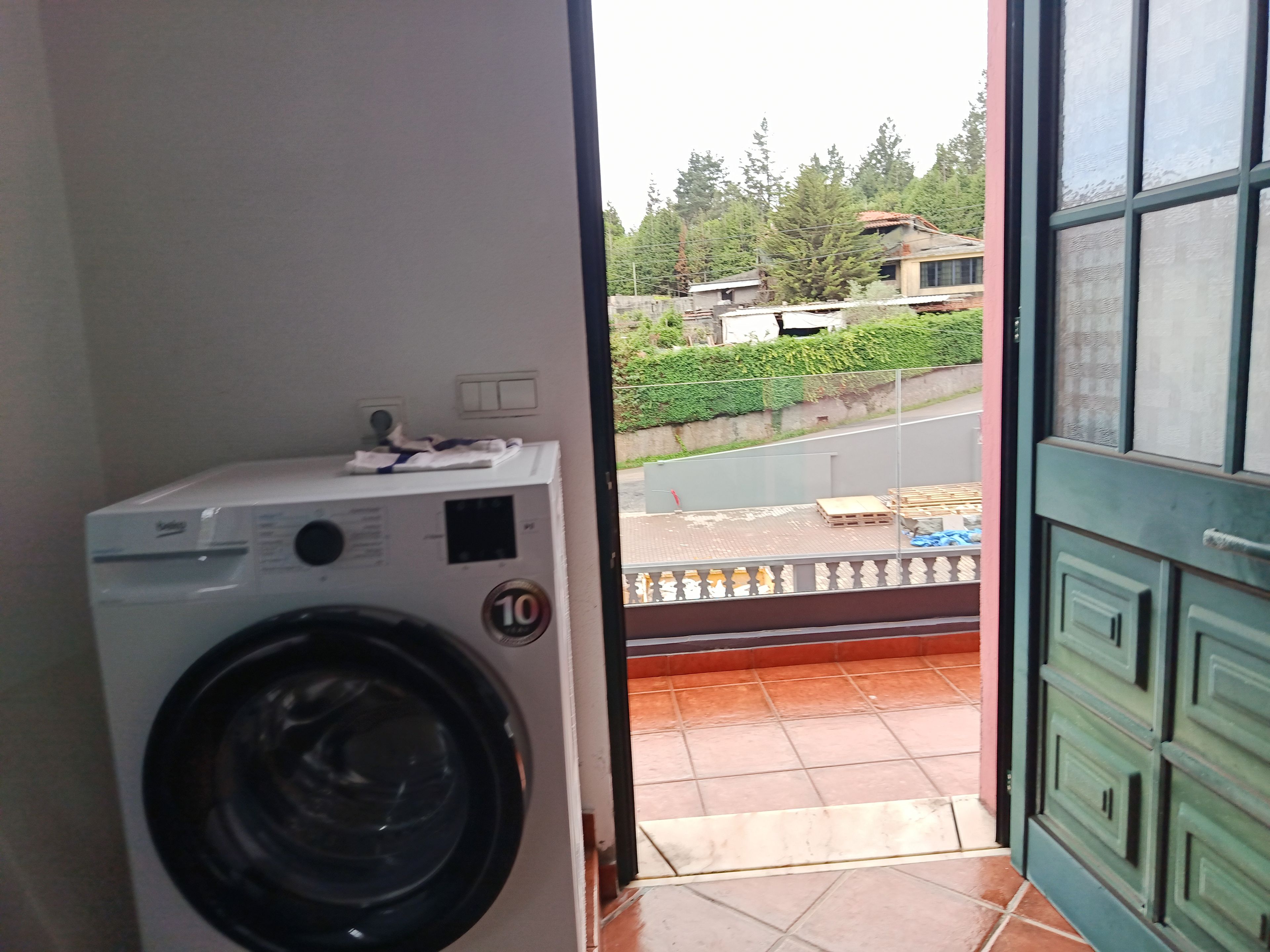 A room with a washing machine, a door leading outside, and a view of a street and greenery.