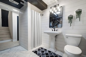 Een badkamer met een wit wastafeltje op een zuil, een toilet, een douche met witte gordijnen en een spiegel met verlichting.