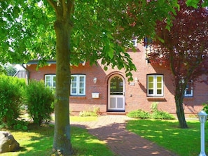 A brick building with a central arched entrance, numbered 28, surrounded by a well-maintained garden and a paved pathway. - Strandstrasse OG (Nieblum)