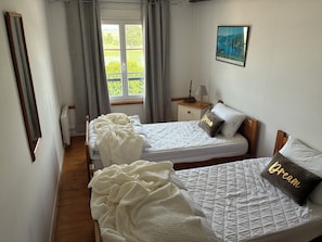 A bedroom with two beds, a window with curtains, a bedside table with a lamp, and a framed picture on the wall. - My beautiful berry (Saint-Valery-sur-Somme)