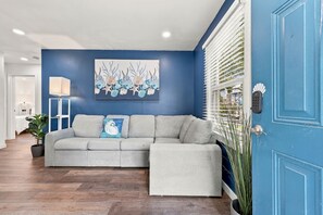 A living room with a grey sectional sofa, a blue accent wall, a painting of sea shells, a lamp, and a door with a keypad. - Sea Shell at Sea Breeze Bungalows 1 Block to Beach (North Myrtle Beach)