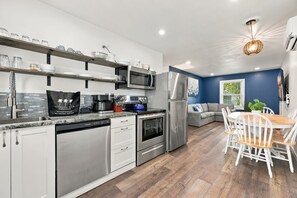 A modern kitchen with stainless steel appliances, a dining area with a wooden table and white chairs, and a blue accent wall. - Sea Shell at Sea Breeze Bungalows 1 Block to Beach (North Myrtle Beach)