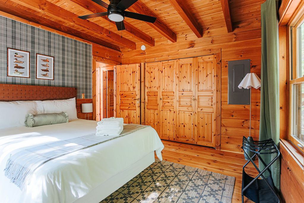 Ein Schlafzimmer einer Holzhütte mit einem Bett, einem Deckenventilator, einem Fenster mit Vorhängen und einer Wand mit gerahmten Bildern.