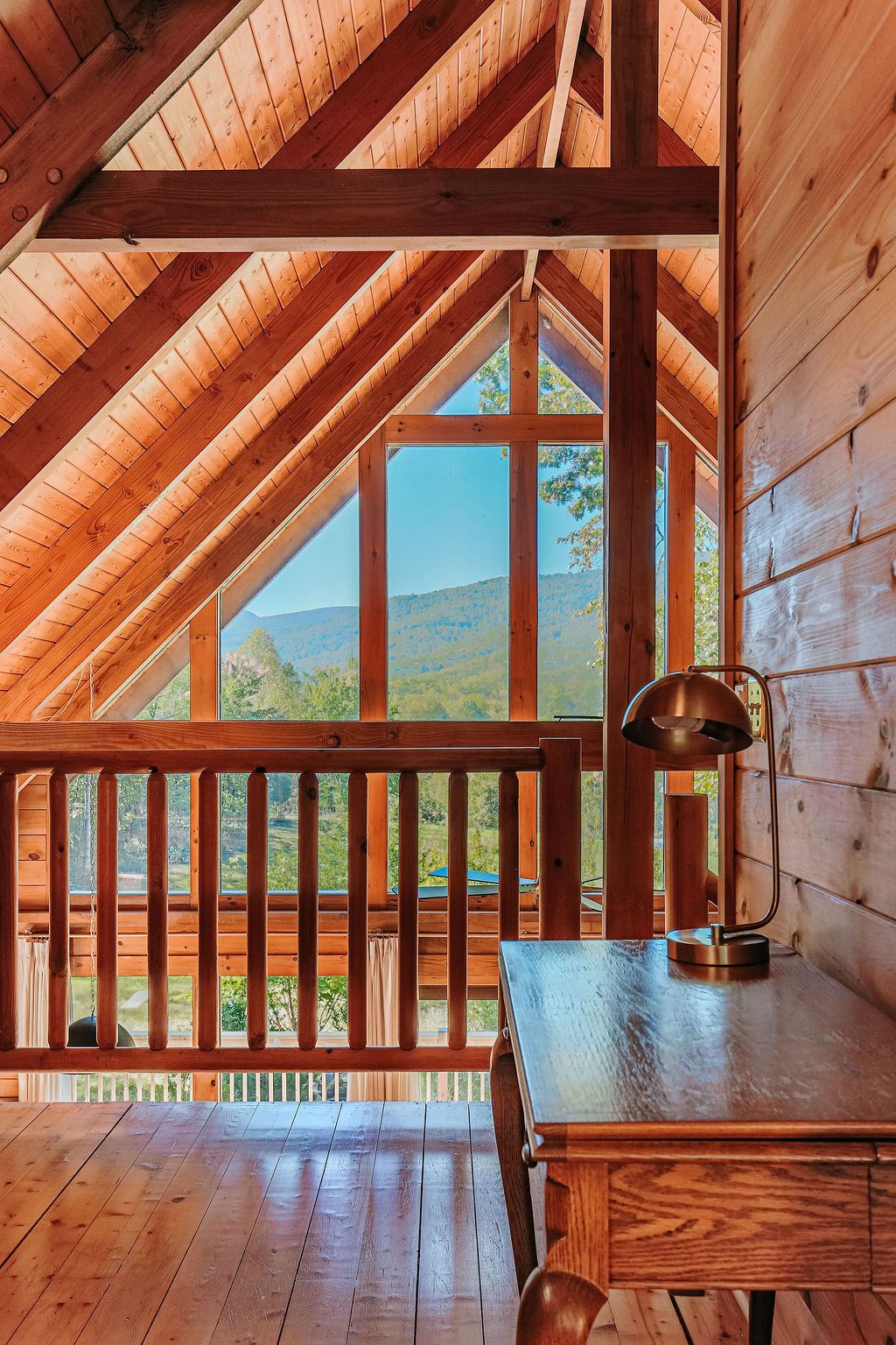 A wooden interior with a sloped ceiling, a wooden desk, and a view of a landscape through large windows.