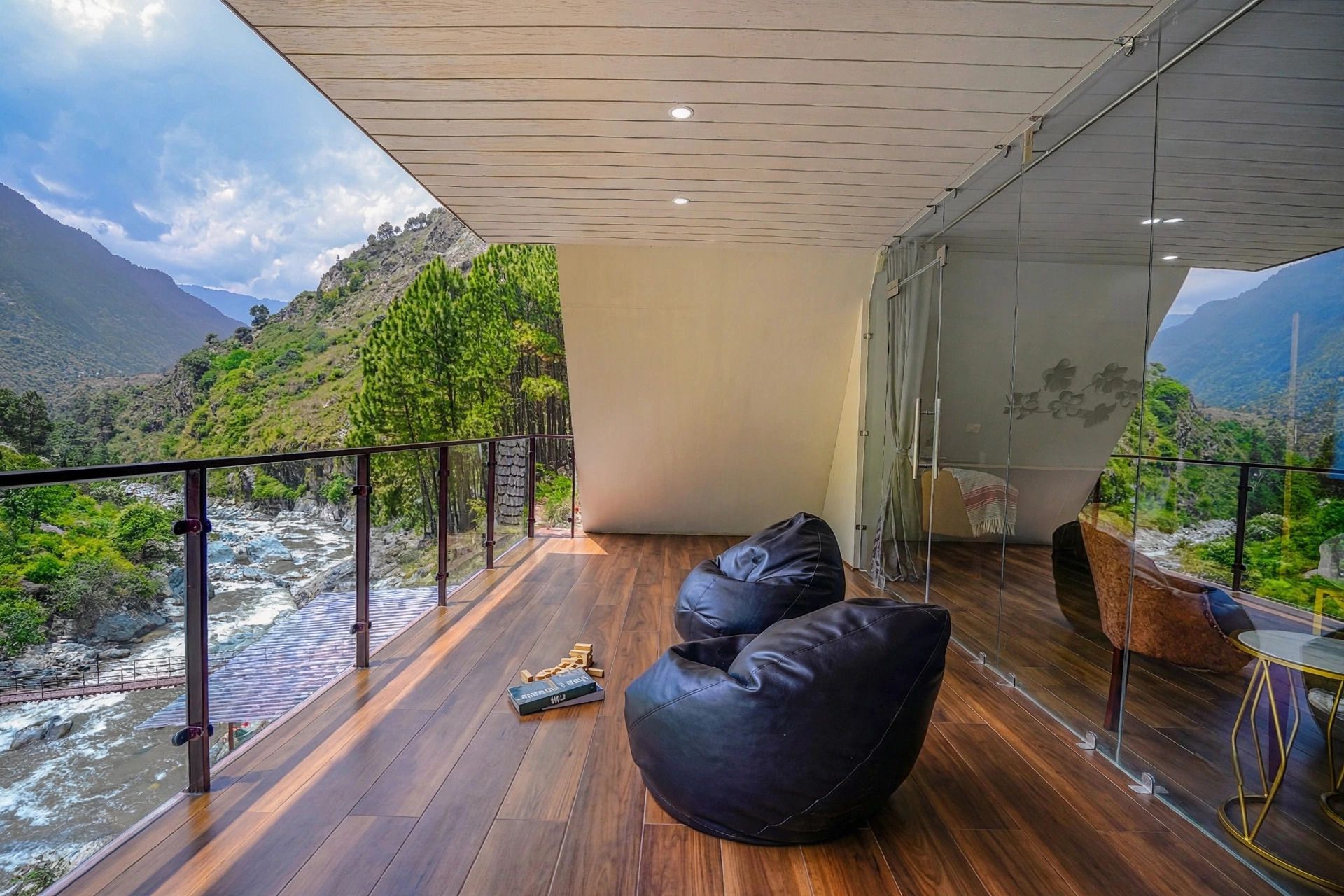Een moderne kamer met een houten vloer, een zwarte relaxfauteuil en een glazen tafel. Een glazen wand biedt uitzicht op een rivier en bergen.