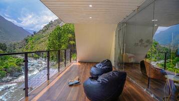 Een moderne kamer met een houten vloer, een zwarte relaxfauteuil en een glazen tafel. Een glazen wand biedt uitzicht op een rivier en bergen.