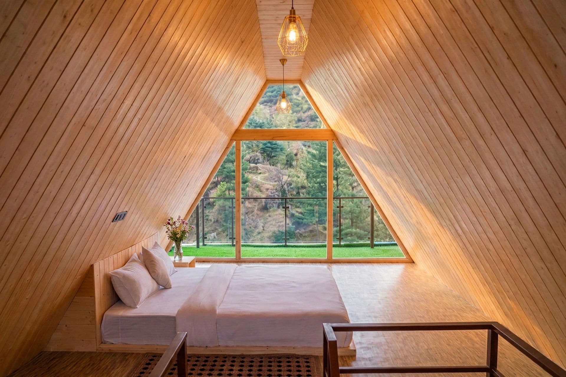 Una habitación acogedora con techo inclinado, una cama, una mesita y vistas a un paisaje verde a través de amplios ventanales.