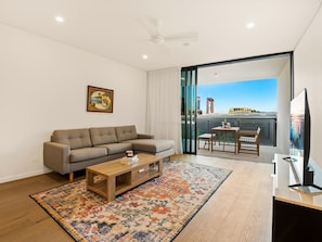 A modern living room with a sofa, coffee table, and a TV. There's a balcony with a view of a cityscape. - 2BR West End Apt w/ Parking Walk to Convention Ctr (West End)