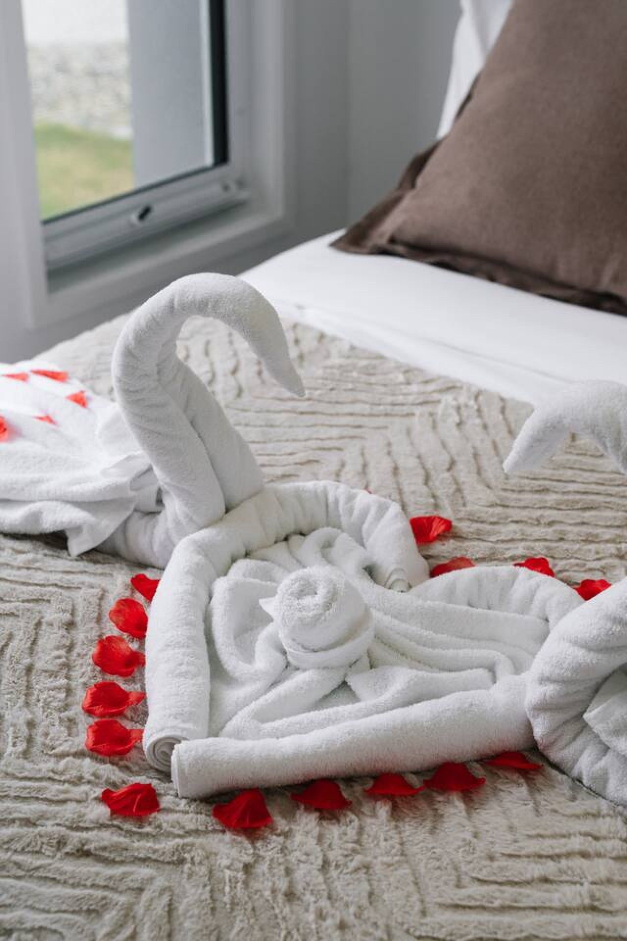 Una cama con un edredón blanco texturizado, una toalla enrollada y pétalos de rosa roja.