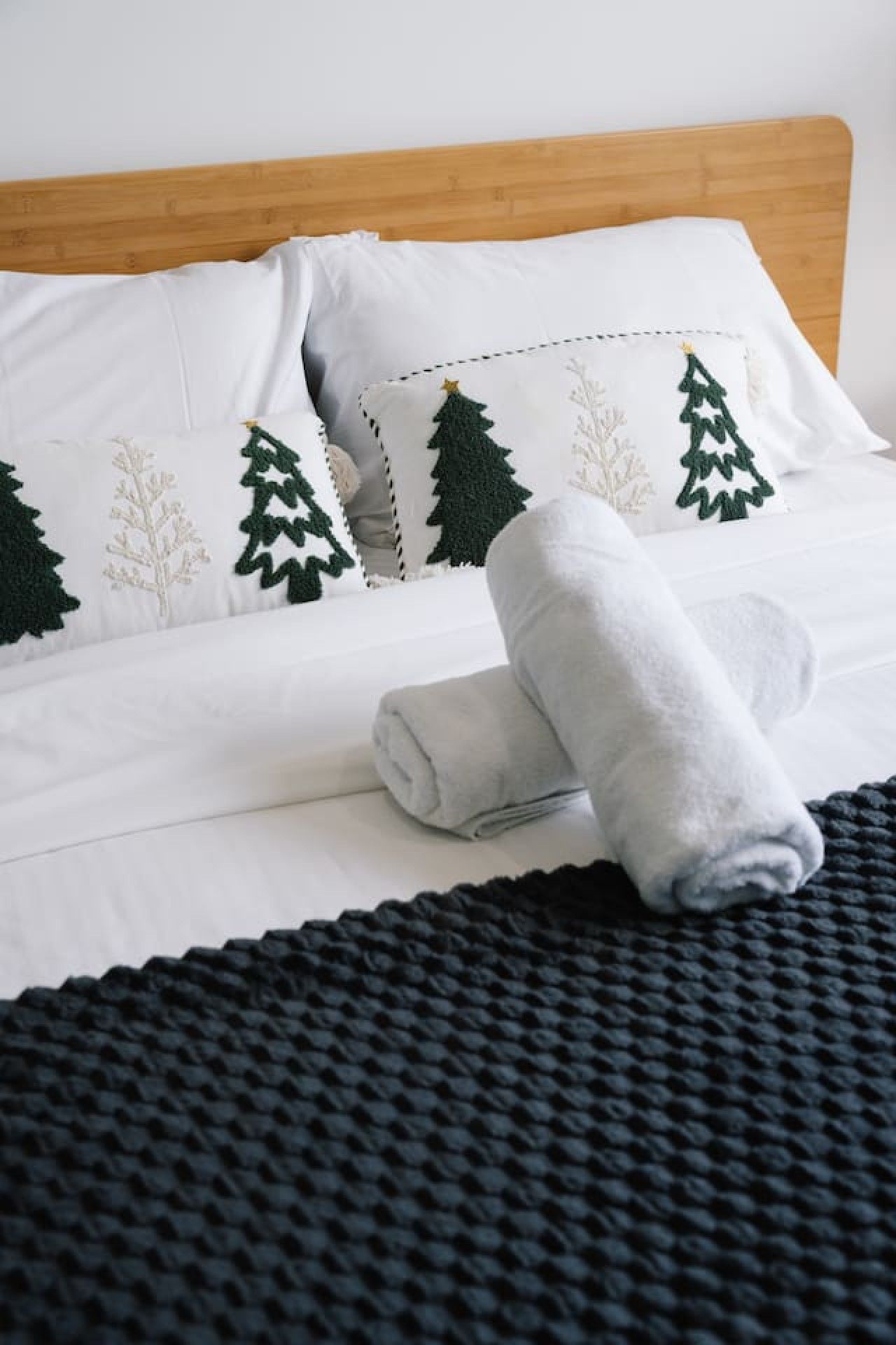 A neatly made bed with white bedding and green tree-shaped pillows, a rolled gray towel, and a black textured blanket.