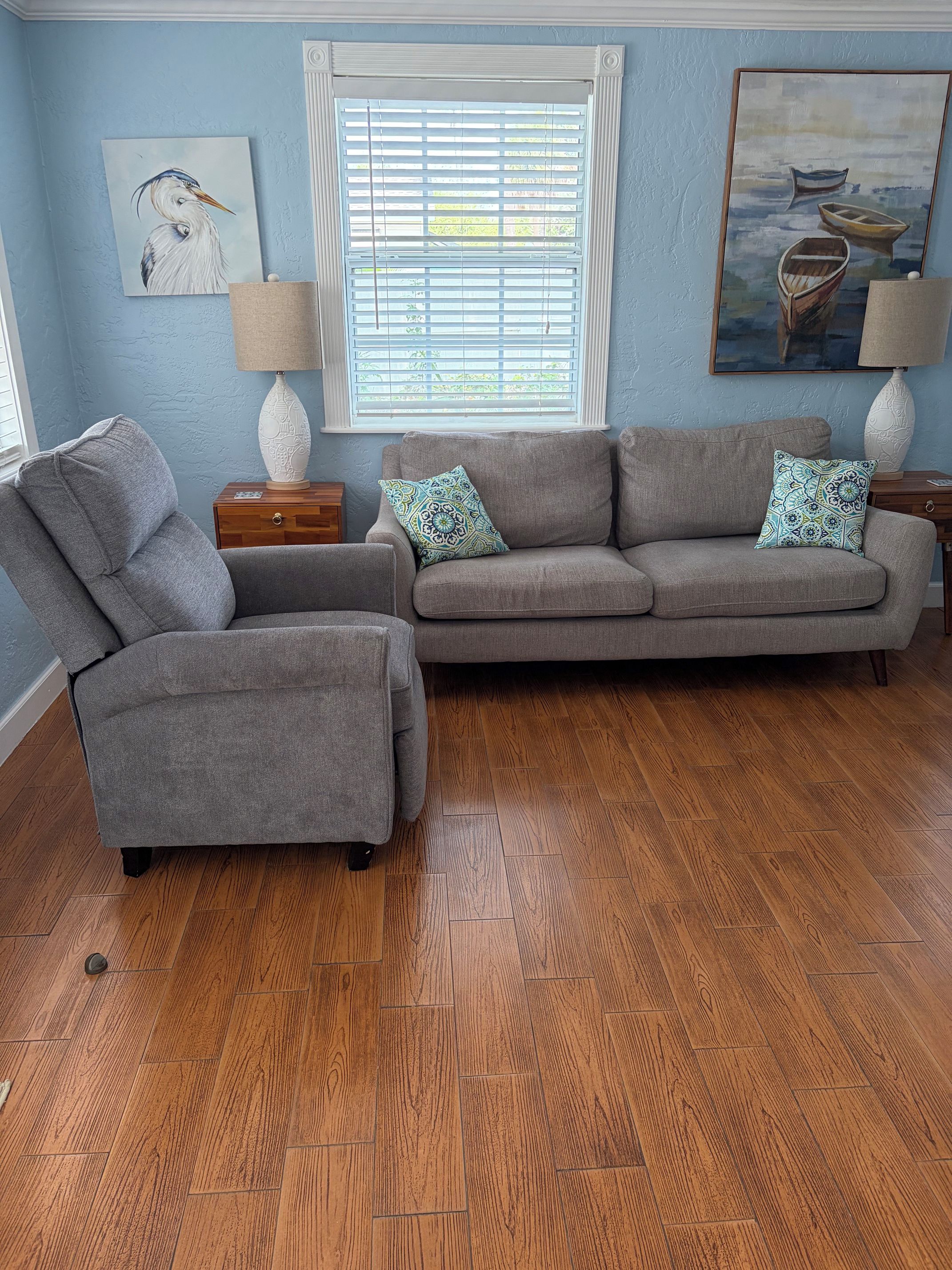 A living room with a grey sofa, a grey armchair, wooden flooring, a window with blinds, and two wall-mounted lamps.