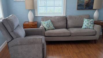 A living room with a grey sofa, a grey armchair, wooden flooring, a window with blinds, and two wall-mounted lamps.