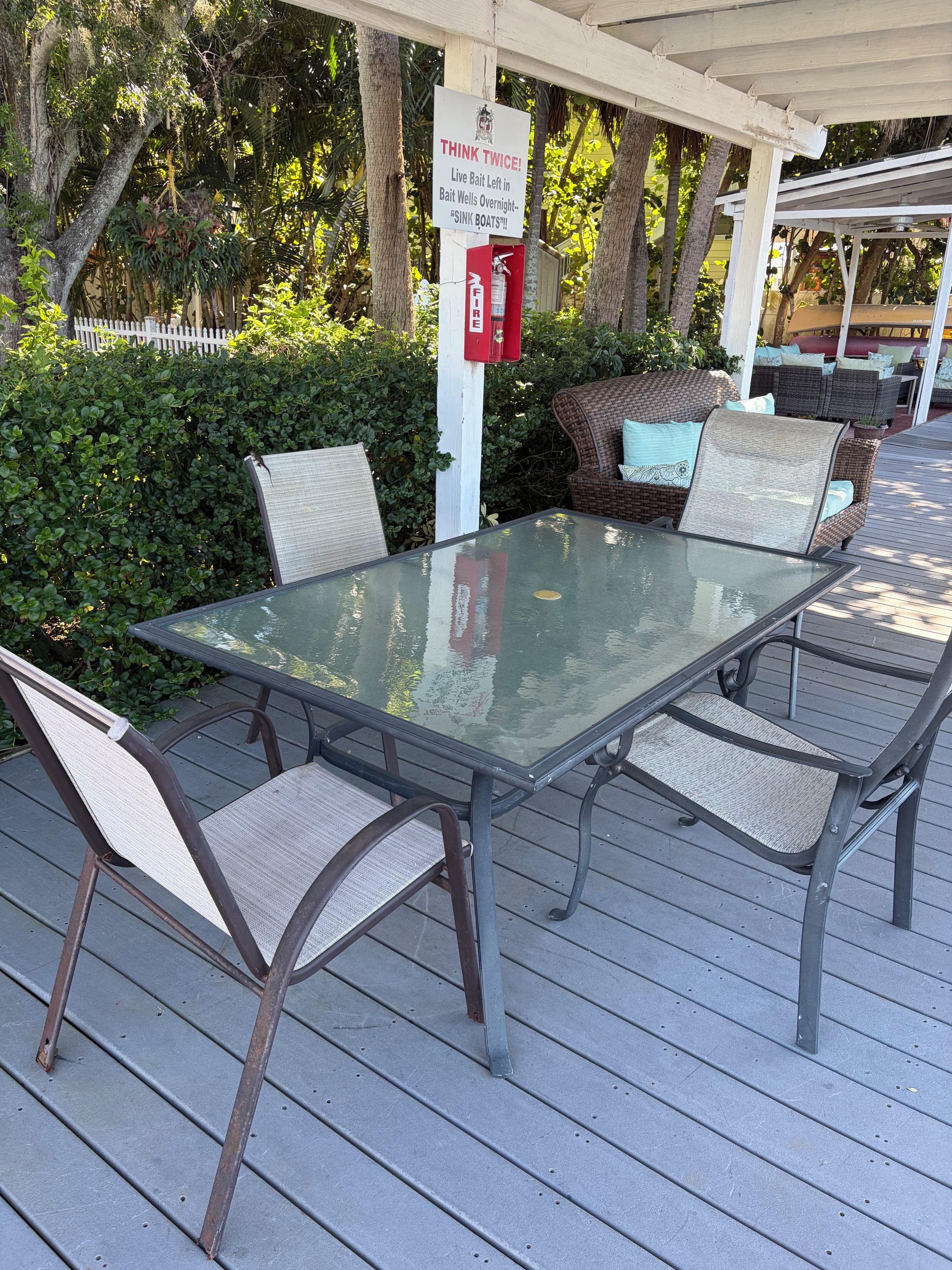Un patio al aire libre con una mesa de vidrio y sillas de mimbre, rodeado por una terraza de madera y follaje.