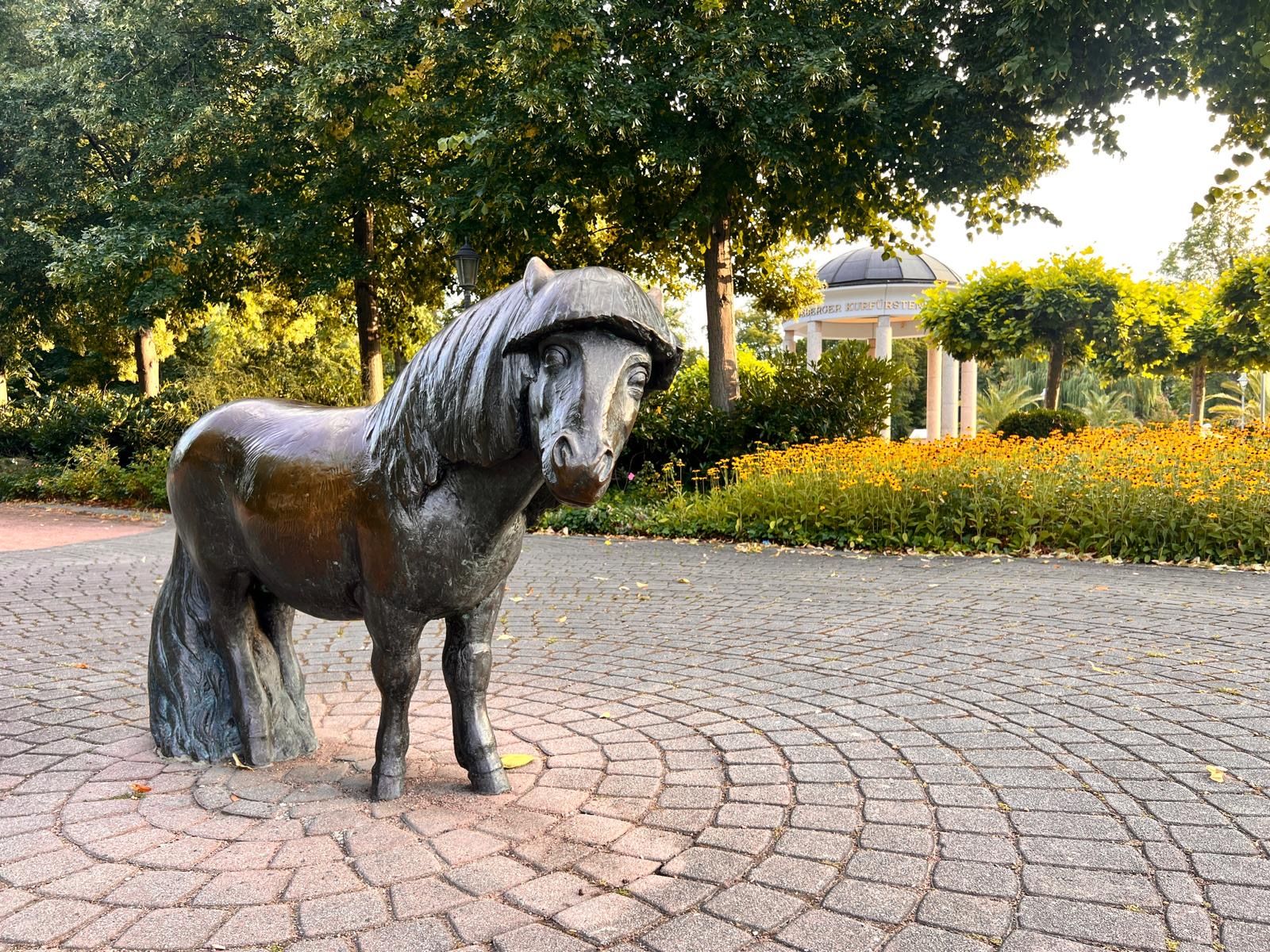 Een bronzen beeld van een paard in een park, met een geplaveid pad, omringd door groen en een gebouw met een koepel op de achtergrond.