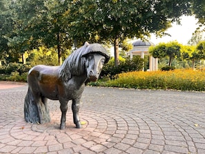 Une statue équestre en bronze se dresse dans un parc, bordée d’une allée pavée, entourée de verdure et avec un bâtiment surmonté d’un dôme en arrière-plan.