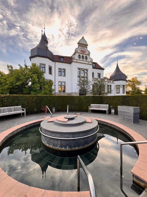 A circular water feature with a central fountain, surrounded by a paved area with benches. In the background, there is a large building with a distinctive architectural style, including a tower and a dome.
