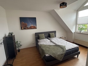 Une chambre à coucher comprenant un lit, une table de chevet, une commode, une fenêtre avec des stores, un luminaire au plafond et un tableau accroché au mur.