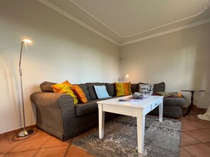 A living room with a sofa, a coffee table, a floor lamp, and a small table with a lamp. - EG - Jaardenhuug (Utersum)