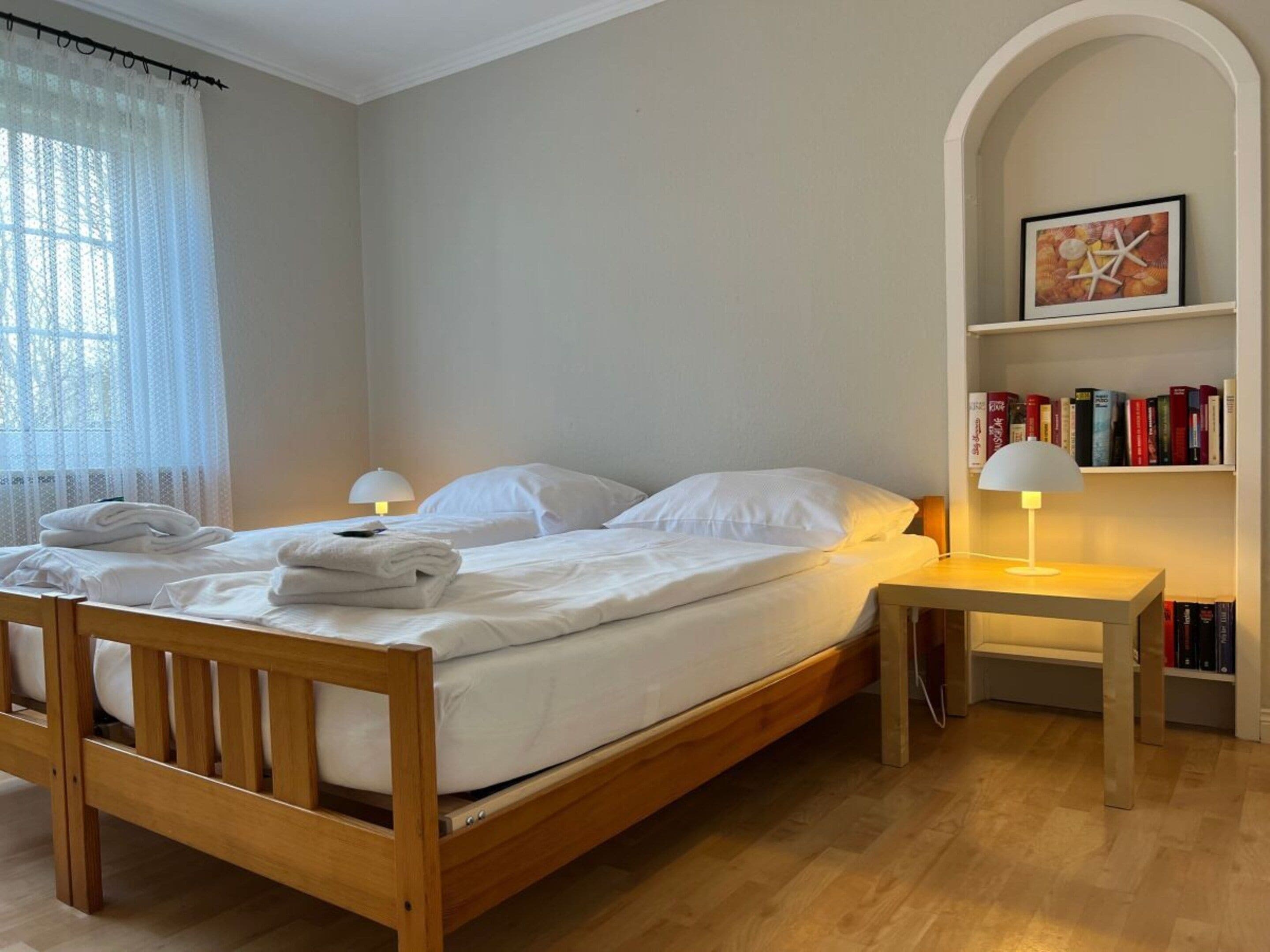 A neatly made bed with white linens, a bedside table with a lamp, and a bookshelf with books.