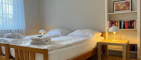 A neatly made bed with white linens, a bedside table with a lamp, and a bookshelf with books.