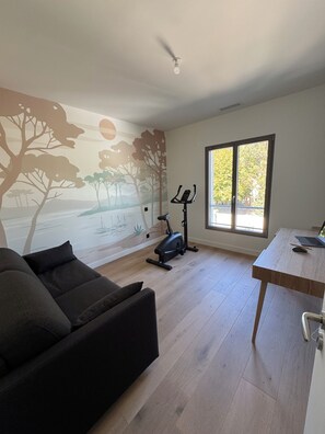 A room with a mural of trees and a sunset, a black exercise bike, a wooden desk with a laptop, and a dark-colored sofa. - Oxéanide: Exceptional villa with a pool (Lacanau)
