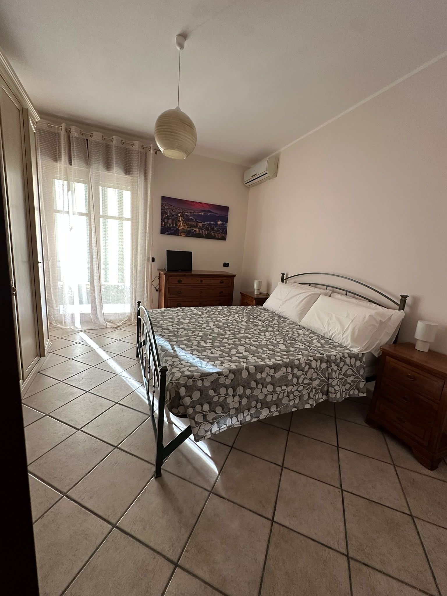 Une chambre à coucher comprenant un lit, une table de chevet, une commode, une fenêtre avec des rideaux et un appareil de climatisation.