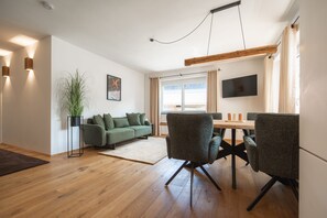 Ett modernt vardagsrum med trägolv, en grön soffa, ett matbord med stolar, en krukväxt och en platt-TV som är monterad på väggen.
