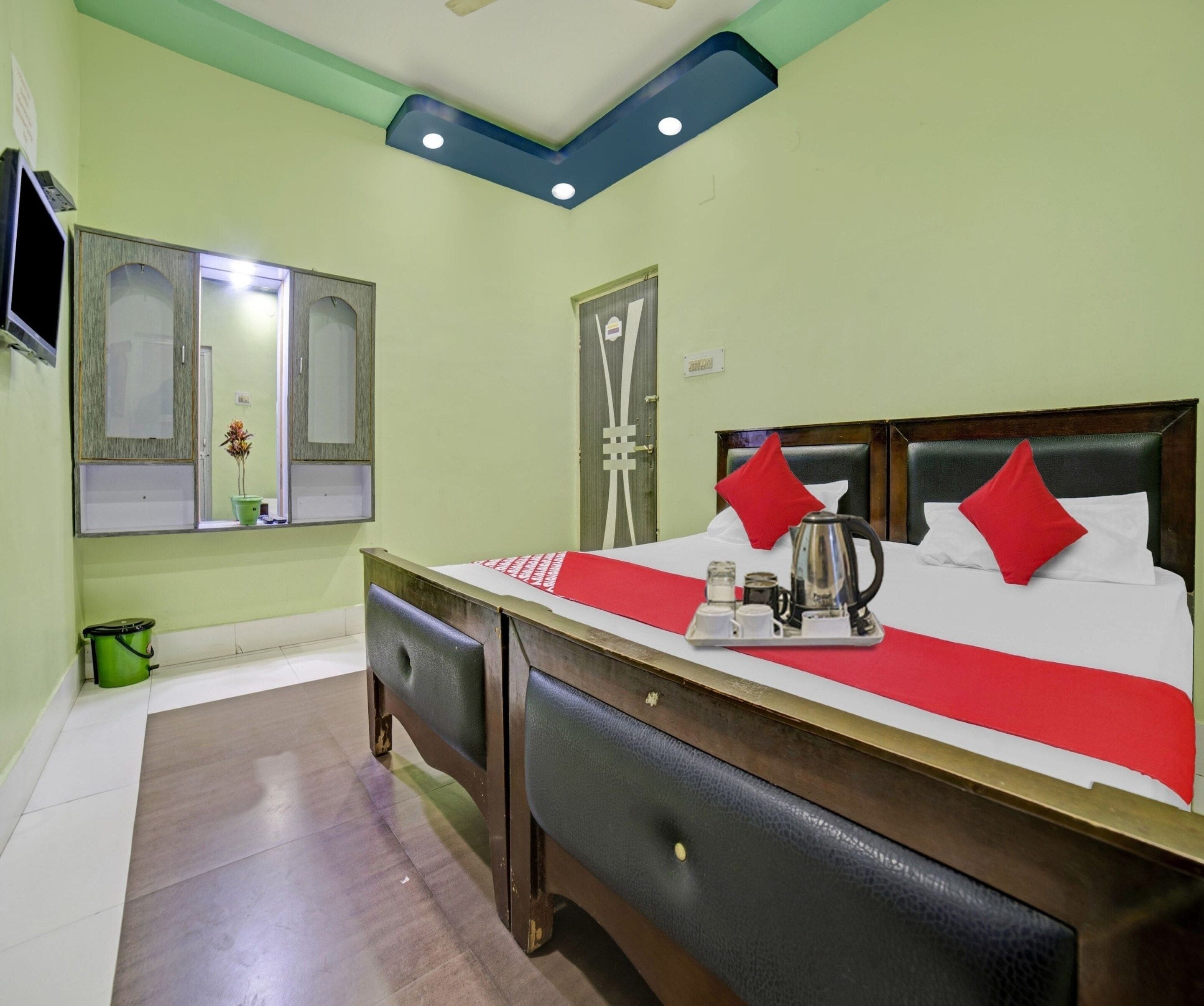 A hotel room with a bed, a bedside table with a kettle, a mirror, and a green bucket.