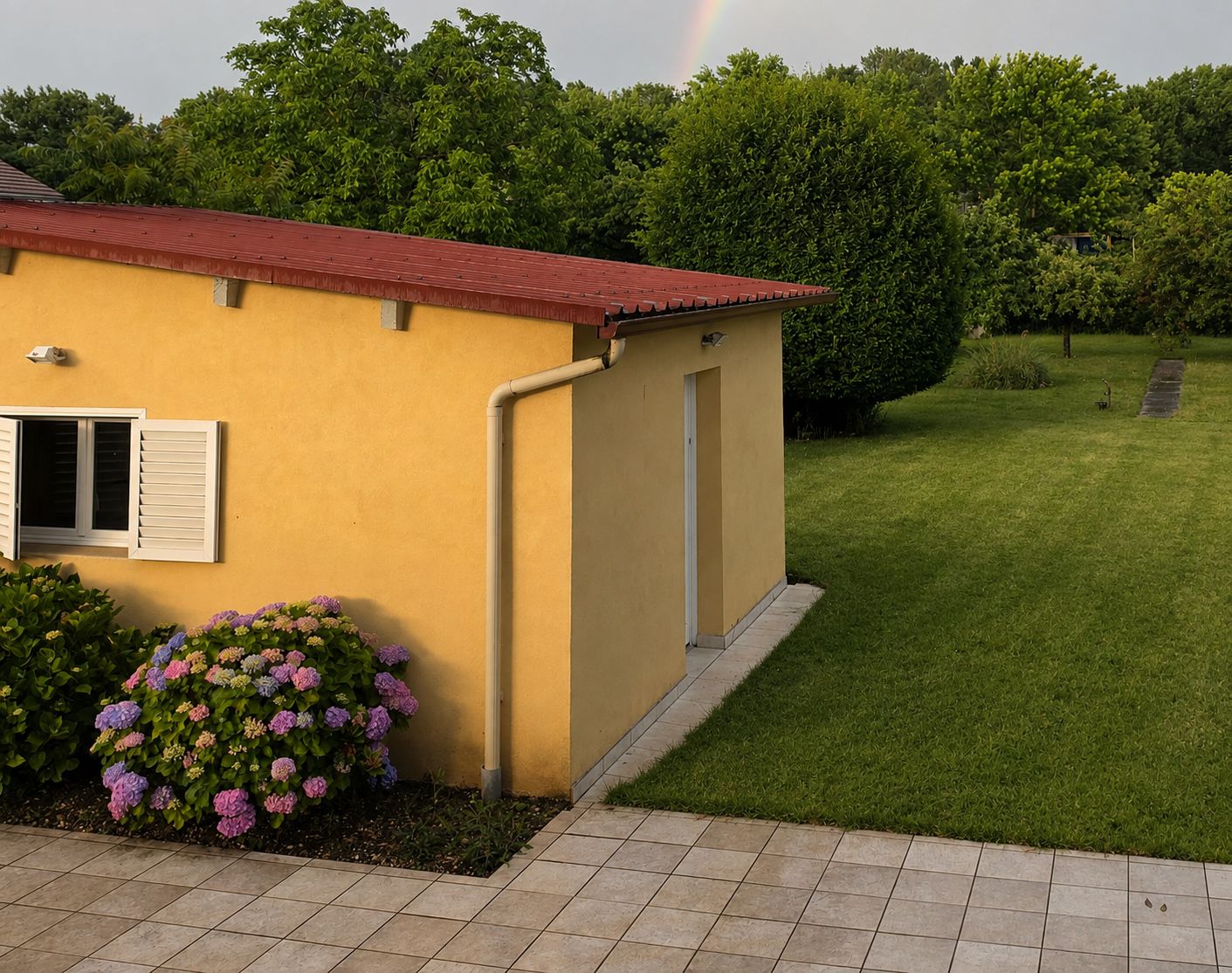 Ein gelbes Gebäude mit rotem Dach, ein kleiner Garten mit Hortensienblüten und einer Rasenfläche.