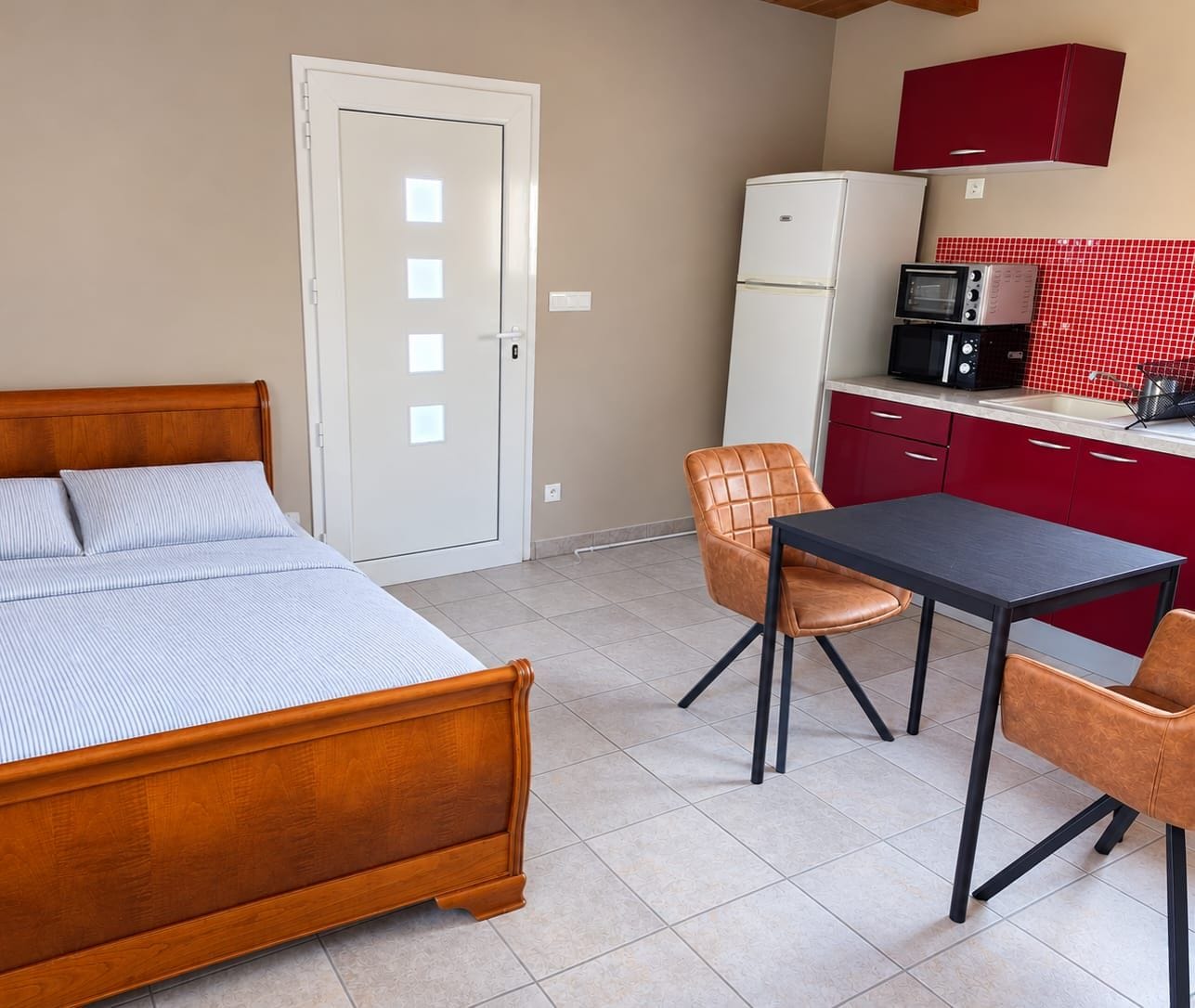 Ein Schlafzimmer mit einem hölzernen Bett, einem kleinen Tisch und zwei Stühlen. Es gibt eine Küchenzeile mit einer Mikrowelle und einem Kühlschrank.