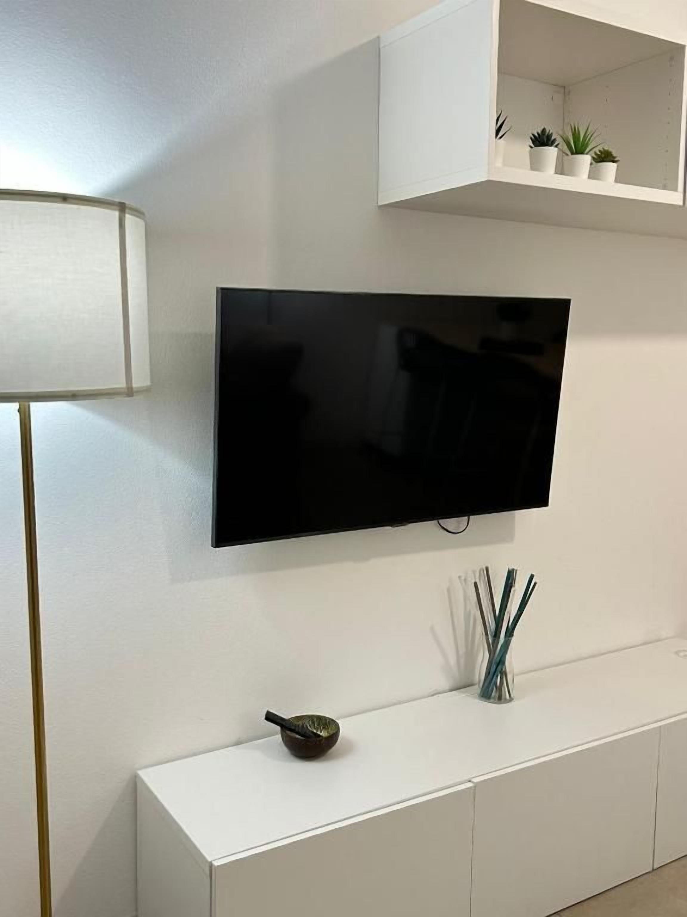 Una sala moderna con una televisión de pantalla plana montada en la pared, una consola blanca con objetos decorativos y un estante blanco con pequeñas plantas.